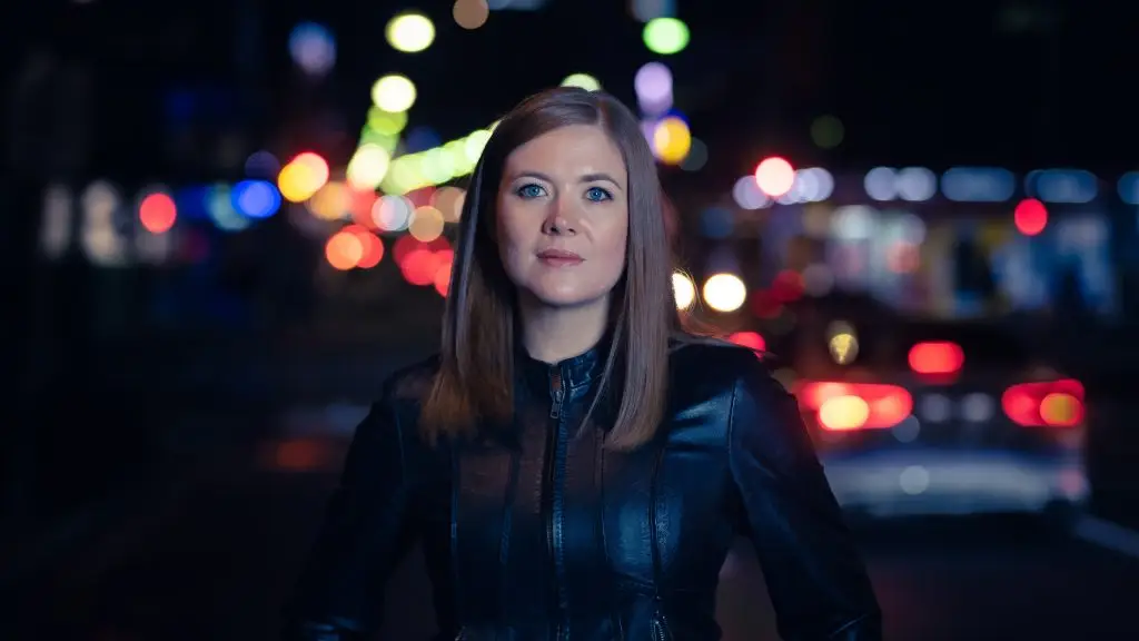 A woman with brown hair stands in front of blurred city lights. She is wearing a black leather jacket and lightly smiling at the camera