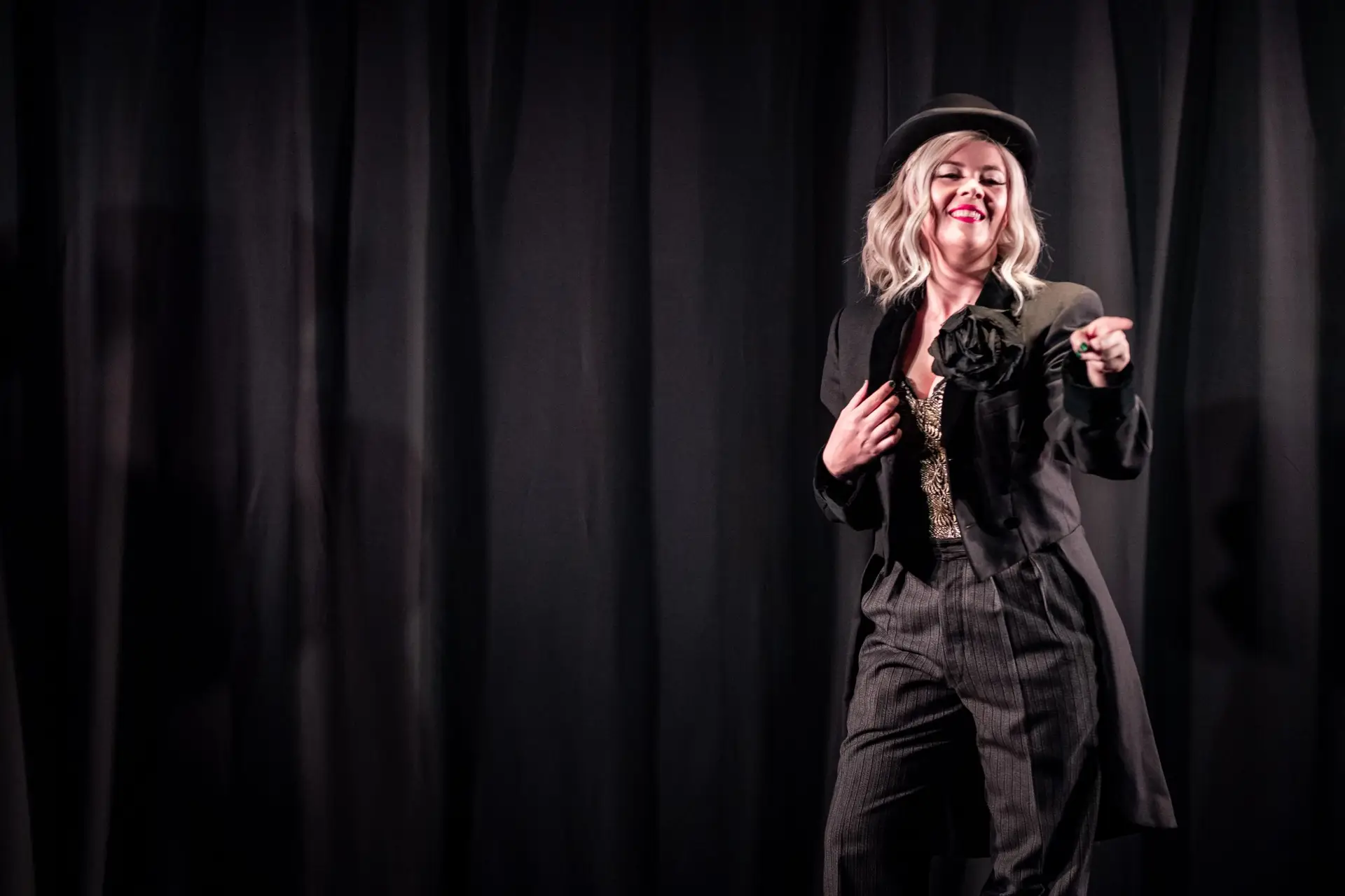 A female performer with white blonde hair is pointing to the audience. She is wearing dark grey trousers and a black coat with tails, and a black hat